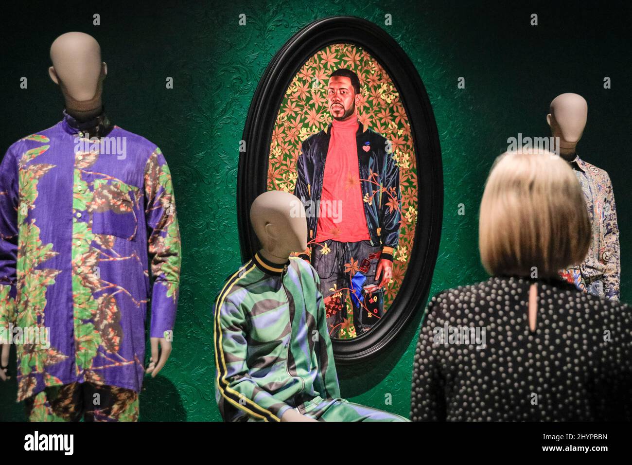 London, Großbritannien. 15. März 2022. EMBARGO bis 16. März, 00,01 Uhr GMT. Mitarbeiter der Galerie mit einer Kollektion von Outfits, darunter Designer aus Afrika und Asien, die lokale Muster und Stoffe verwenden, mit Kehinde Wileys Porträt von Alexander Cassatt in der Mitte. Die neue Ausstellung der V&A „Moisting masculinities: The Art of Menswear“ präsentiert rund 100 Looks von legendären Designern und aufstrebenden Stars wie Harris Reed, Gucci, Grace Wales Bonner und Raf Simons sowie 100 Kunstwerke in einer Vielzahl von Medien. Kredit: Imageplotter/Alamy Live Nachrichten Stockfoto