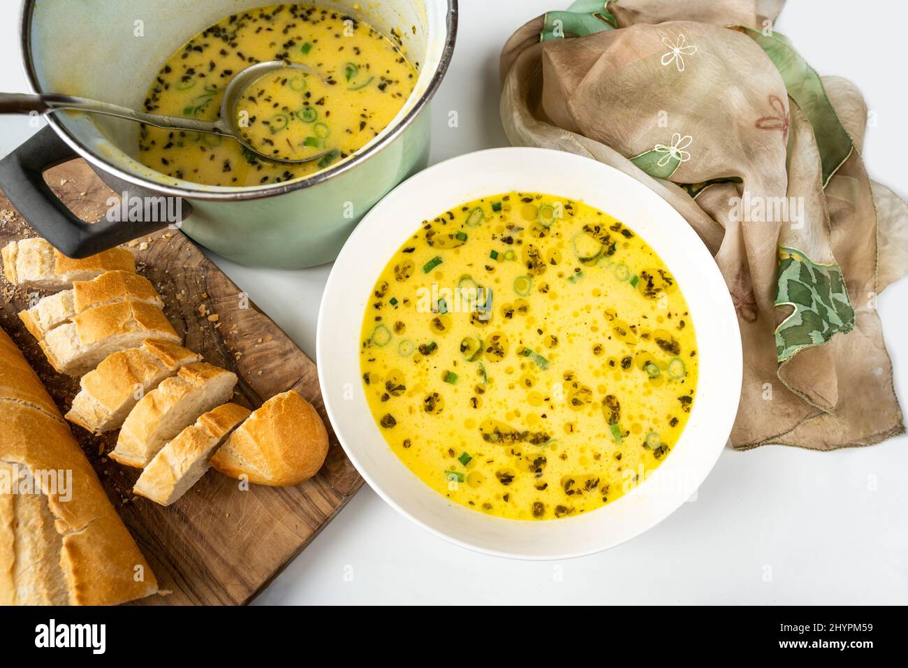 Käsesuppe mit Orangano-Kraut, grünem Zwiebelaufstrich und Öl auf weißem Teller, Brotscheiben, Schal und Topf mit Suppe und Schöpfkelle. Stockfoto