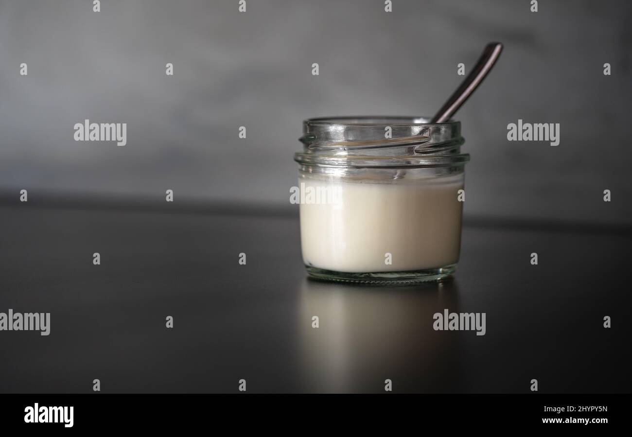 Bio hausgemachter Joghurt in einem kleinen kurzen Glasbehälter mit einem kleinen Löffel, schwarzer Tisch mit Spiegelung des Joghurtbehälters, verschwommener Hintergrund o Stockfoto