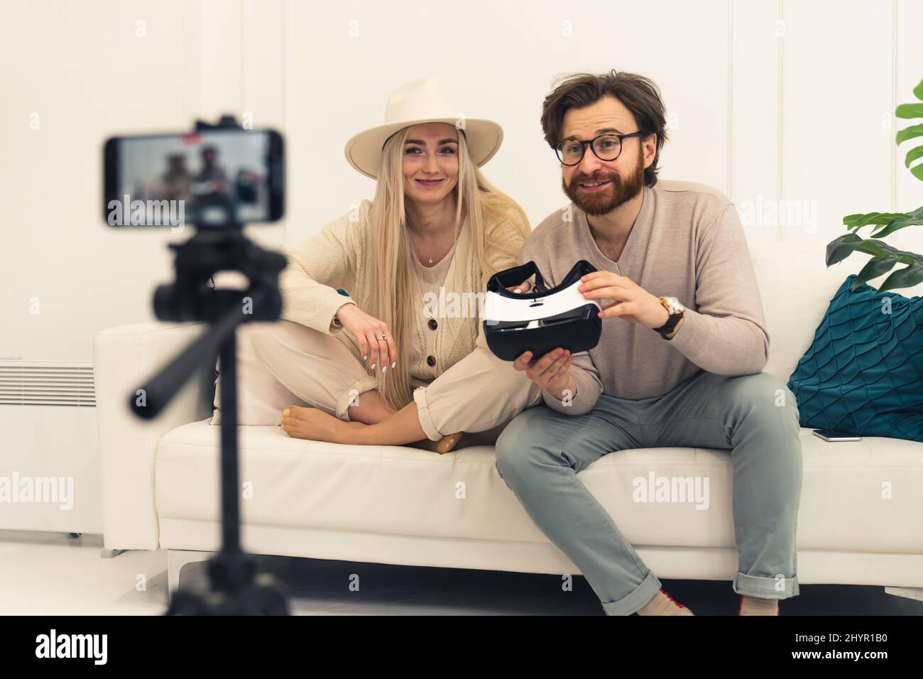 Niedliches kaukasisches Paar, das ein Video mit einem Smartphone auf einer Halterung aufzeichnet, während es eine Brille für die virtuelle Realität zeigt. Mittlere Weitwinkelaufnahme, weißer Hintergrund. Hochwertige Fotos Stockfoto