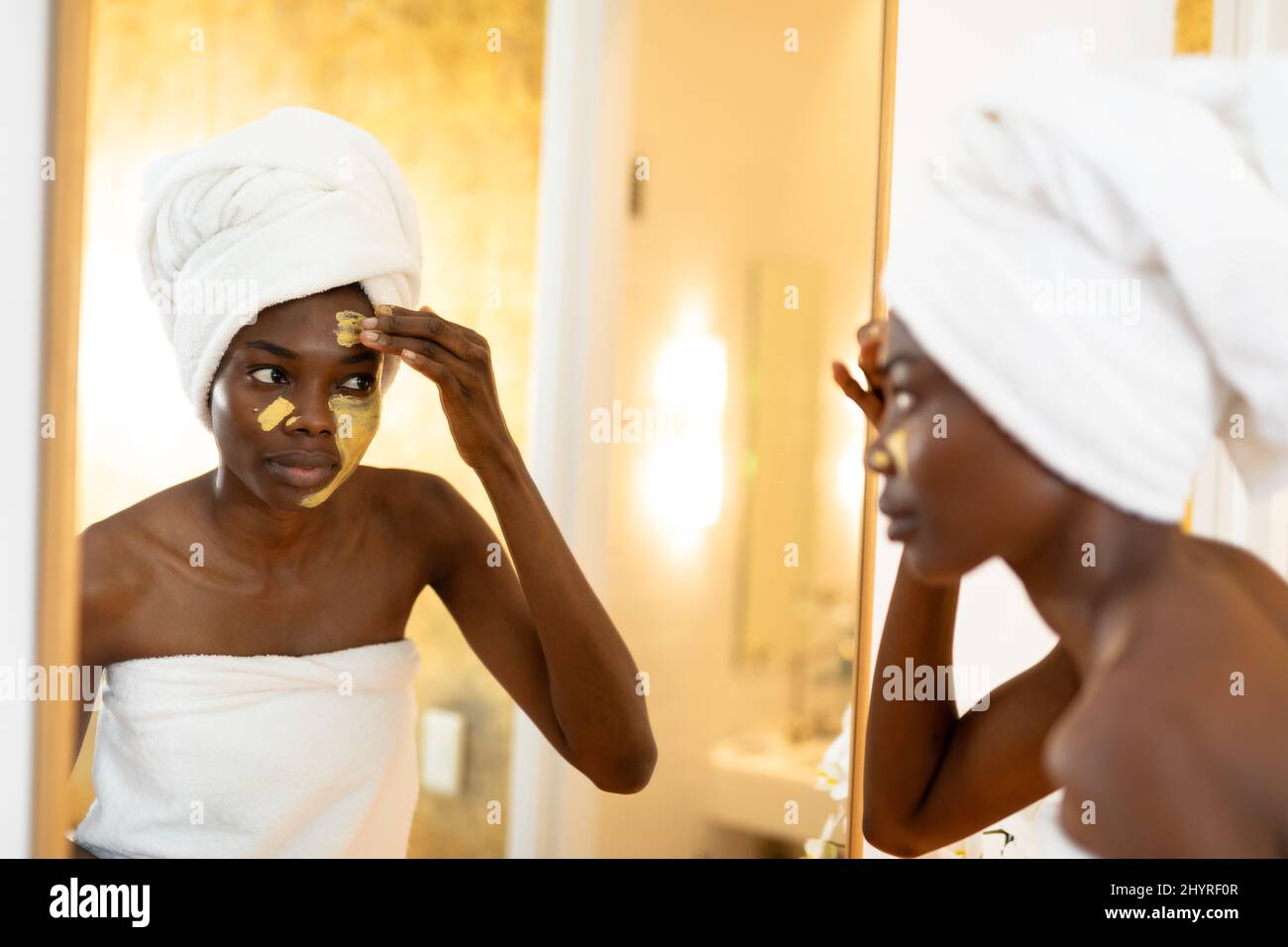 afroamerikanische junge Frau, die Gesichtsmaske aufführt, während sie Spiegelreflexion im Badezimmer betrachtet Stockfoto