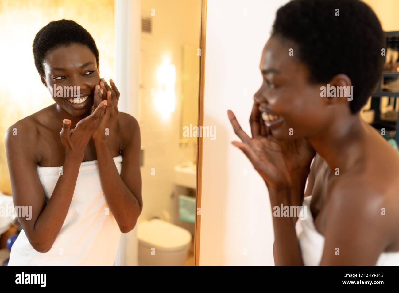 Lächelnde afroamerikanische Frau, die Creme anwendet, während sie auf Spiegelreflexe im Badezimmer schaut Stockfoto