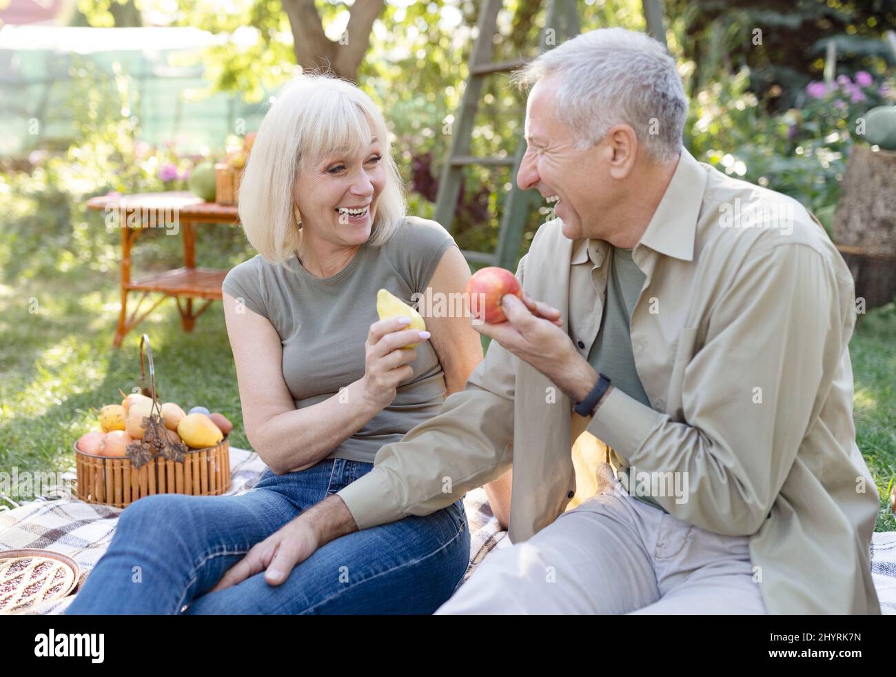 Lachende ältere Ehepartner sitzen auf Feldgras und essen Früchte, reden und lächeln, ruhen sich im Garten aus Stockfoto