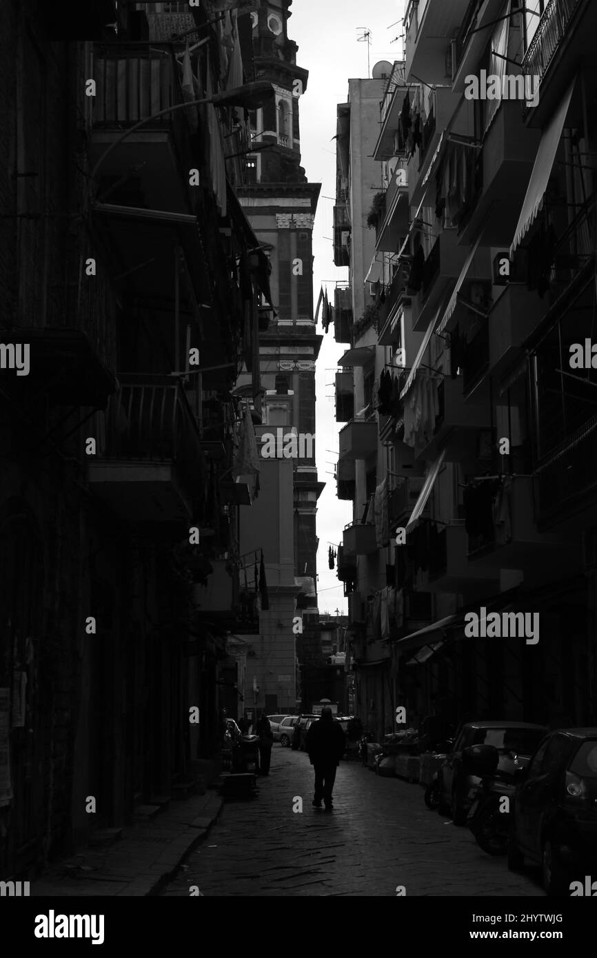 Der Mensch geht in Neapel, Italien, durch eine von der Sonne beleuchtete schmale Straße, während er von überall auf den Balkonen beobachtet werden kann. Stockfoto