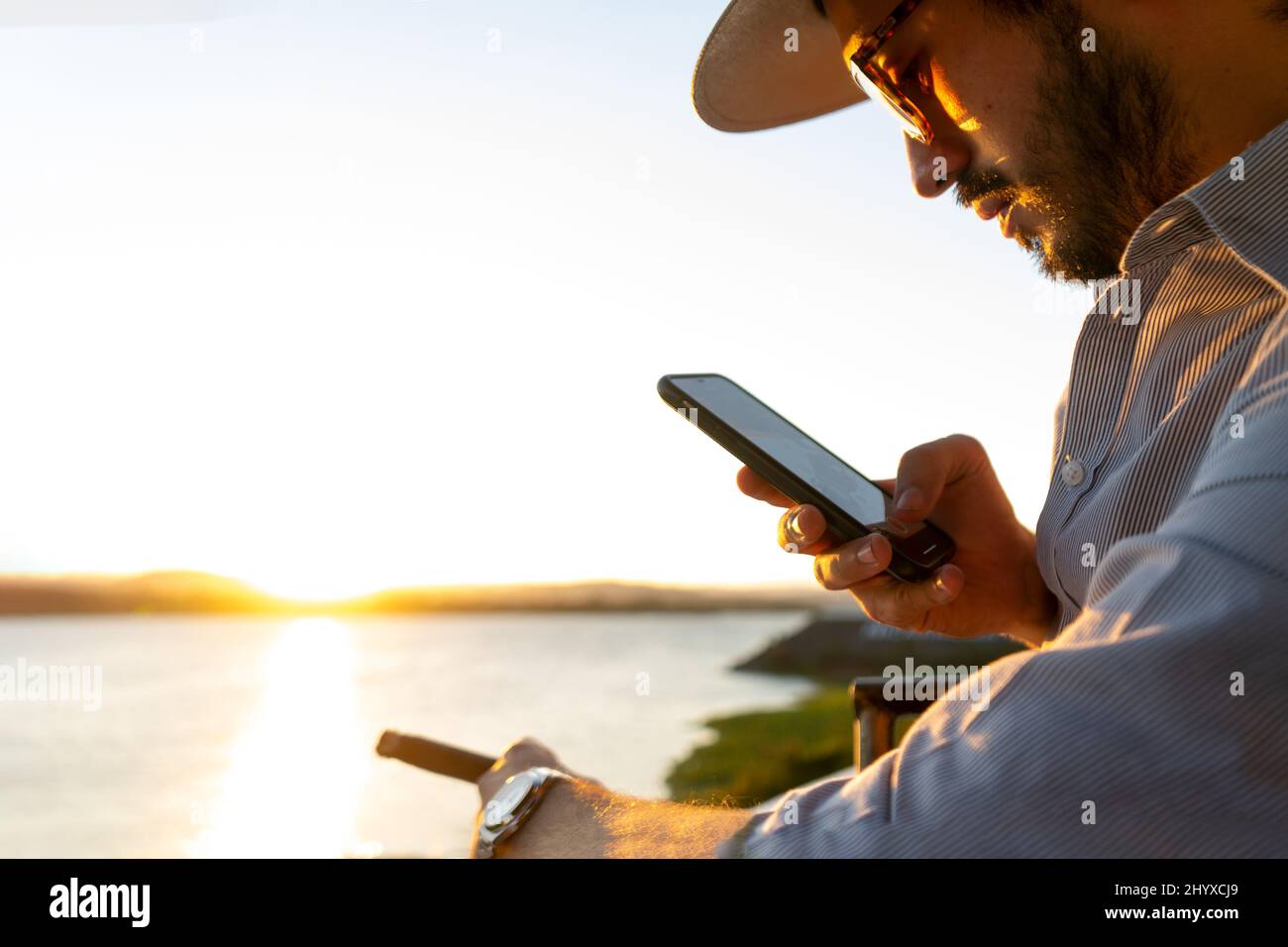 Hipster an der Bar, der ein Foto mit seinem Handy von einer Zigarre während des Sonnenuntergangs an einer privaten Küste machte Stockfoto