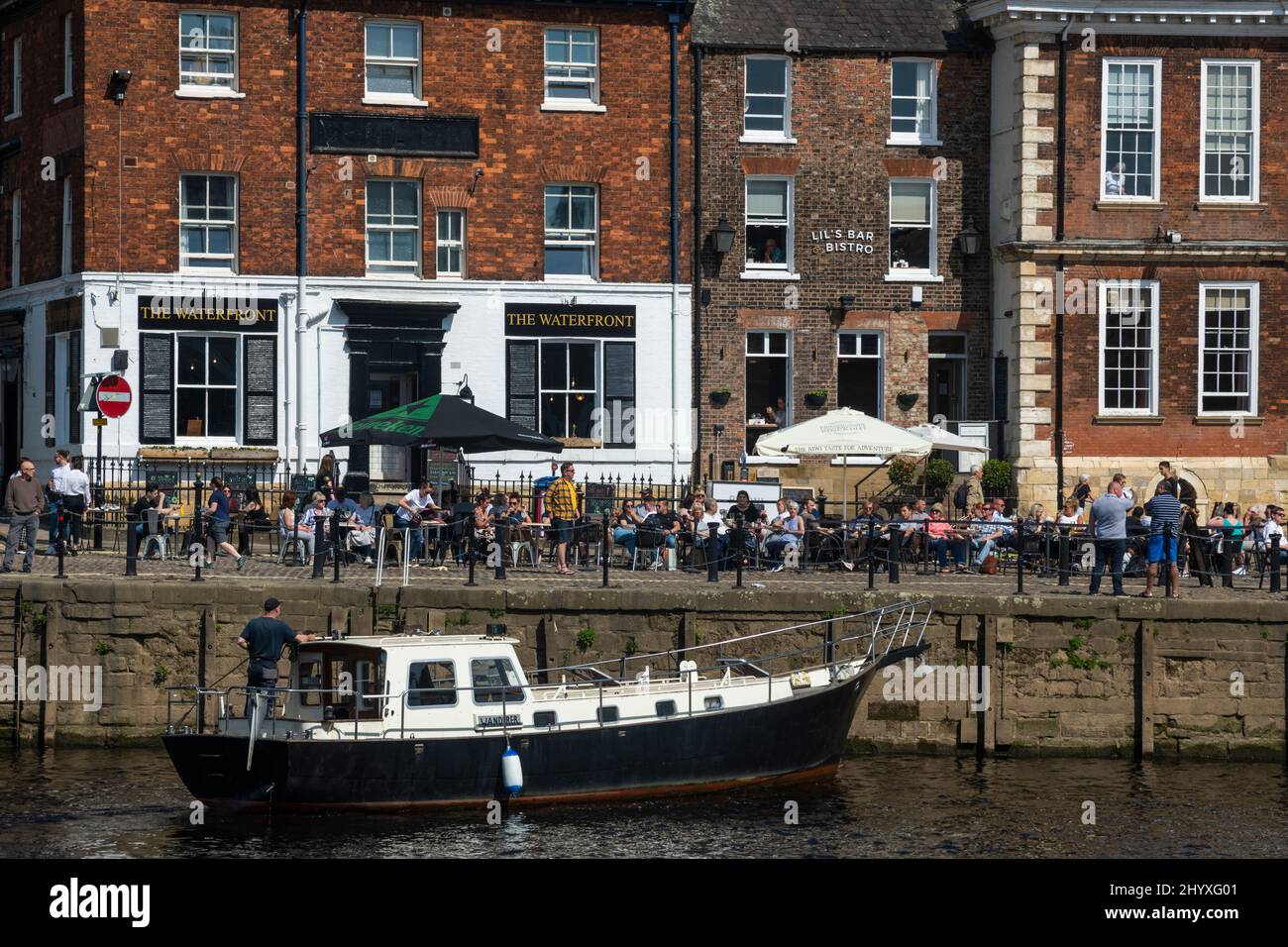 Gäste trinken, essen in belebten Cafés am Flussufer und segeln auf einem Freizeitboot - der malerische sonnige River Ouse, King's Staith, York, North Yorkshire, England. Stockfoto