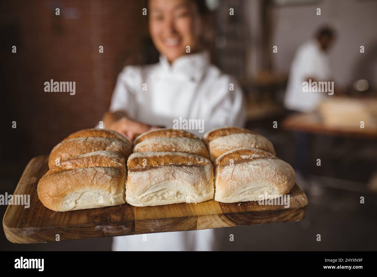 Lächelnde asiatische Bäckerin mittlerer Erwachsenen, die frisch gebackenes Brot auf dem Servierbrett zeigt Stockfoto