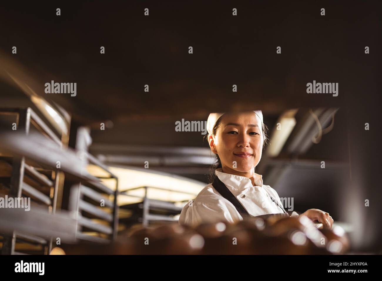 Lächelnde asiatische mittlere Erwachsene weibliche Bäckerin, die geflochtene Brottablett im Regal hält Stockfoto
