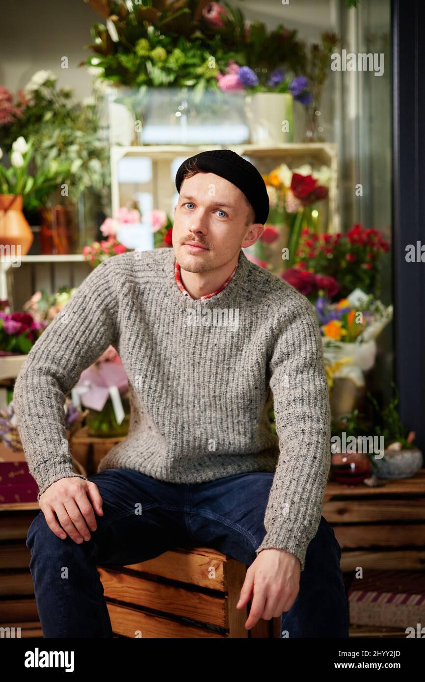 Erfolgreiche stilvolle Unternehmer kaukasischen männlichen Floristen trägt Pullover, Beanie und Brille im Blumenladen. Junger hübscher Gewächshausbesitzer oder Gärtner, der Blumen und Blumensträuße im Geschäft verkauft Stockfoto