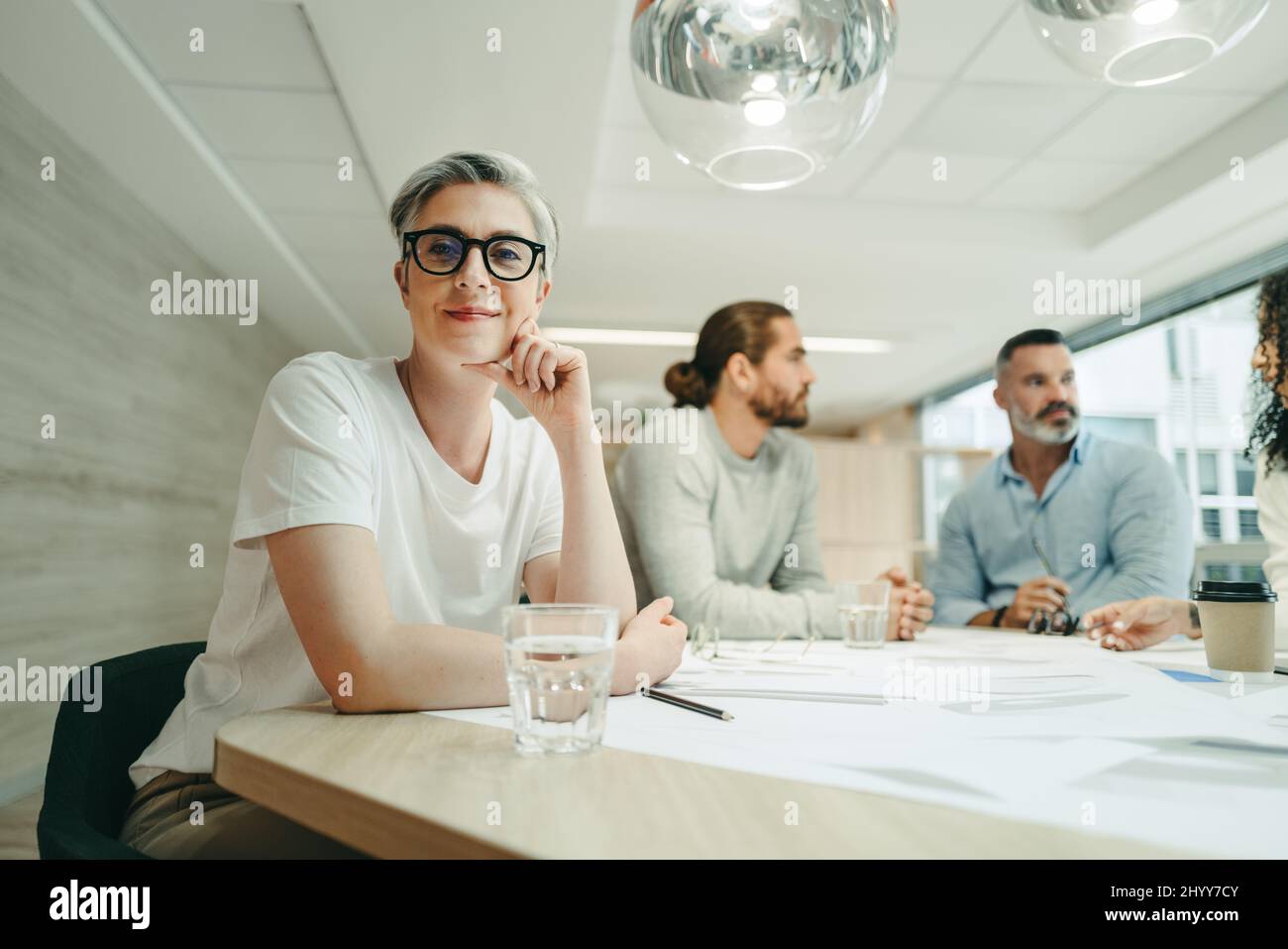 Weibliche Design-Expertin, die die Kamera anschaut, während sie in einem Meeting mit ihrem Team sitzt. Gruppe kreativer Geschäftsleute, die während der Arbeit Brainstorming durchführen Stockfoto