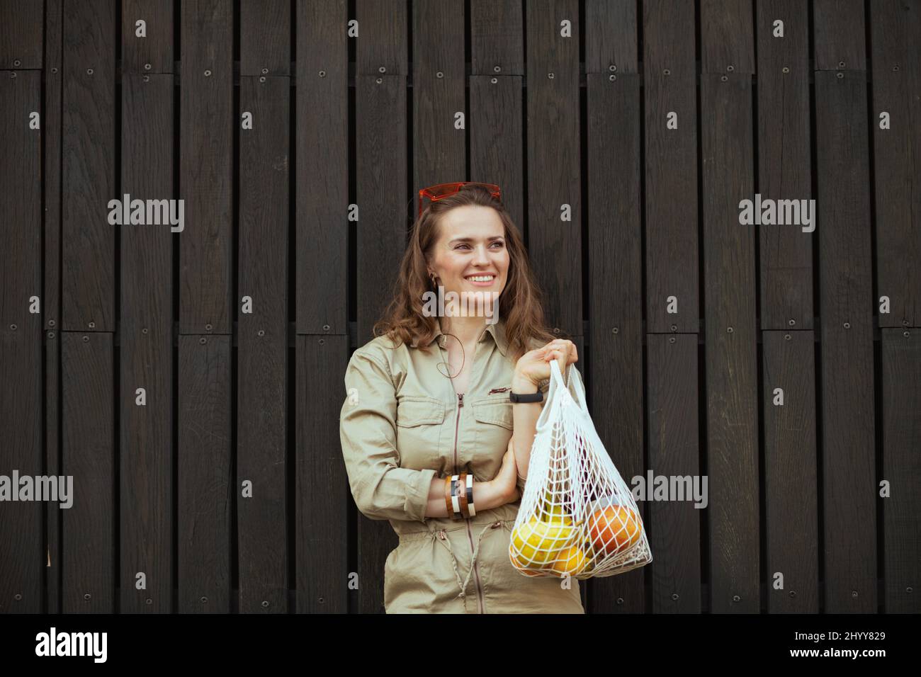 Fröhliche trendige Frau im Overall mit Saitenbeutel gegen schwarze Holzwand im Freien in der Stadt. Stockfoto