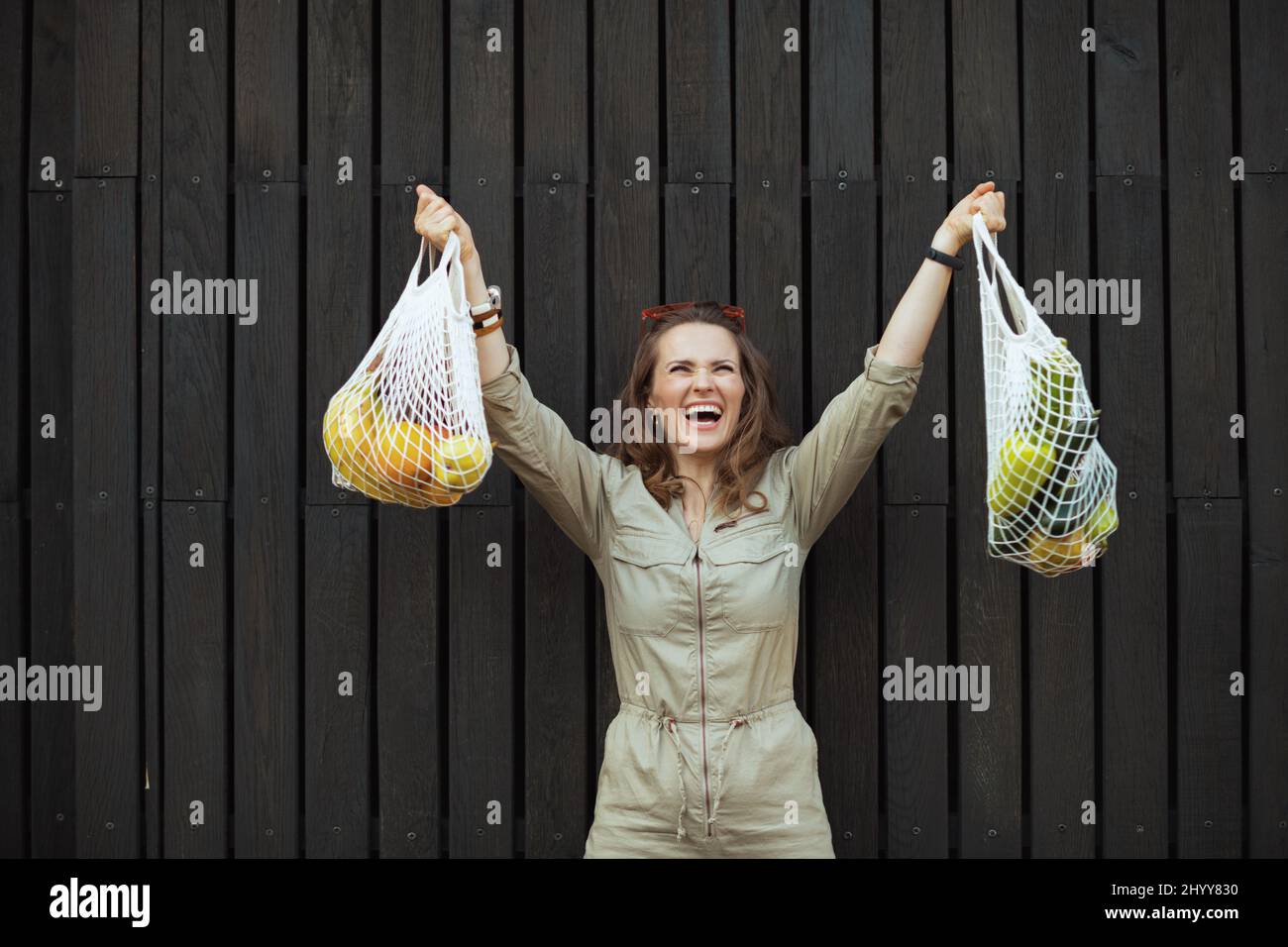 Glückliche elegante Frau im Overall mit Saitenbeutel gegen schwarze Holzwand im Freien in der Stadt. Stockfoto