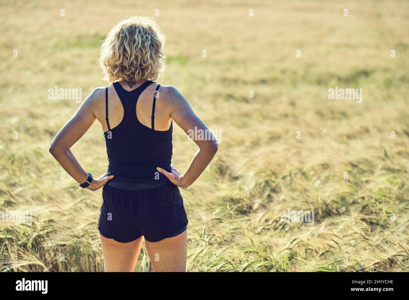 Back Portrait einer jungen reifen kaukasischen fit Frau mit blonden Haaren in Sportswear Outdoor in einer Getreidefarmlandschaft. Lifestyle-Konzept. Stockfoto