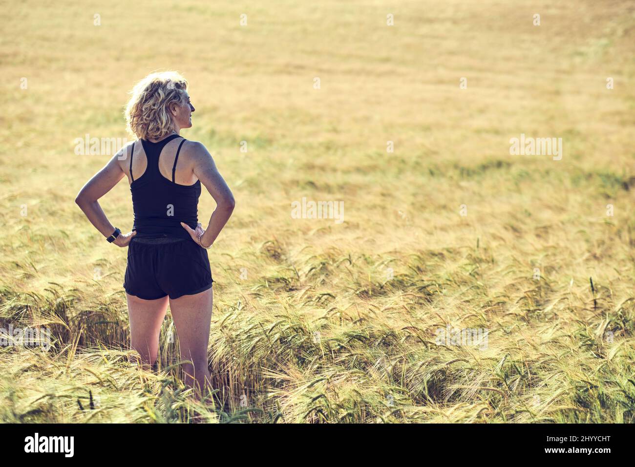 Back Portrait einer jungen reifen kaukasischen fit Frau mit blonden Haaren in Sportswear Outdoor in einer Getreidefarmlandschaft. Lifestyle-Konzept. Stockfoto