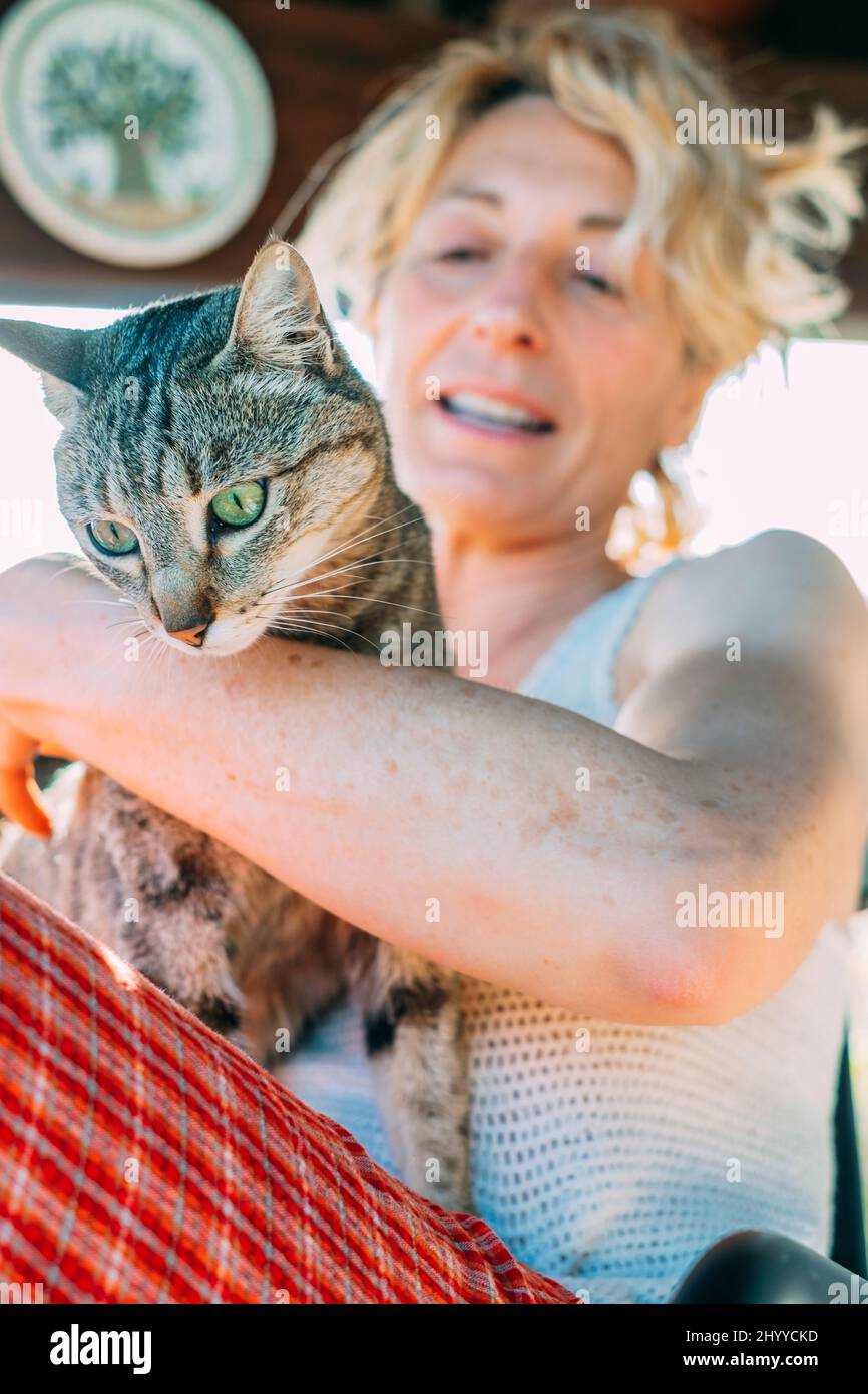 Porträt einer jungen reifen kaukasischen blonden Frau, die mit ihrer Katze zu Hause auf der Veranda lächelt und in einem Schaukelstuhl sitzt. Lifestyle-Konzept. Stockfoto