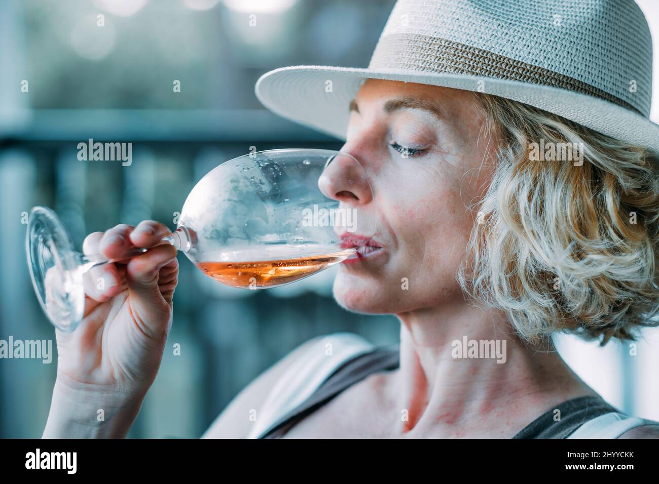 Porträt einer reifen kaukasischen blonden jungen Frau im Alter von 50s Jahren, die einen Hut trägt und ein Glas Wein auf der Veranda eines Landhauses verkostet. Lifestyle c Stockfoto