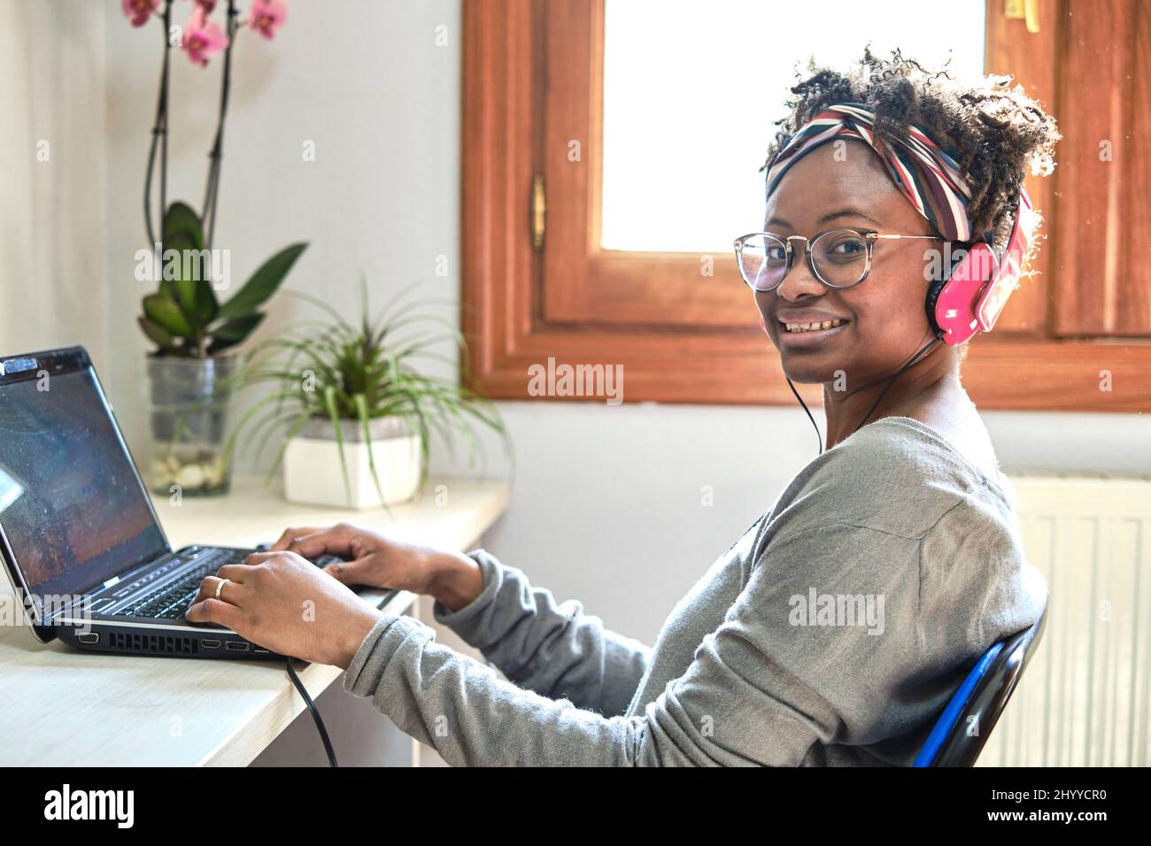 Junge schwarze Frau mit Afro-Frisur und einem Computer und Musik hören mit Kopfhörern in einem Haus. Lifestyle-Konzept. Stockfoto