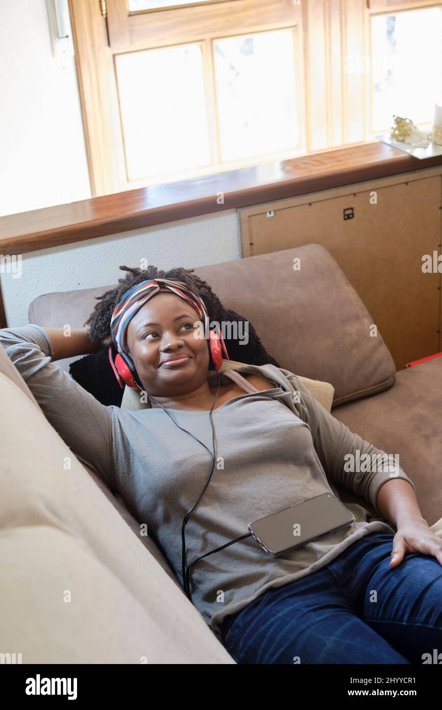 Porträt einer jungen schwarzen Frau, die mit Kopfhörern im Innenbereich Musik hört und auf einem Sofa liegt. Lifestyle-Konzept. Stockfoto
