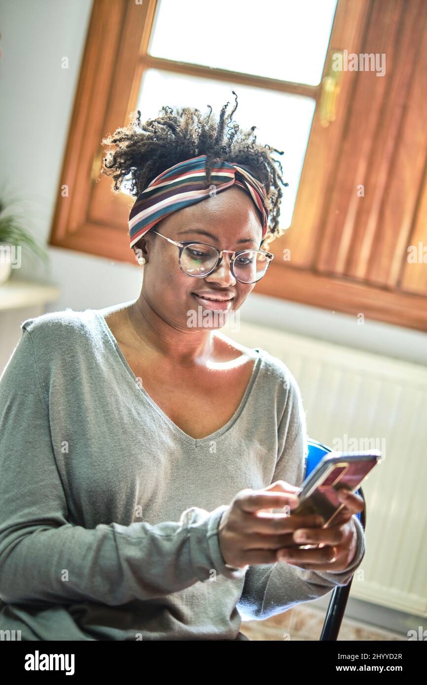 Junge schwarze Frau mit Afro-Frisur und Brille mit Mobiltelefon in einem Haus. Lifestyle-Konzept. Stockfoto
