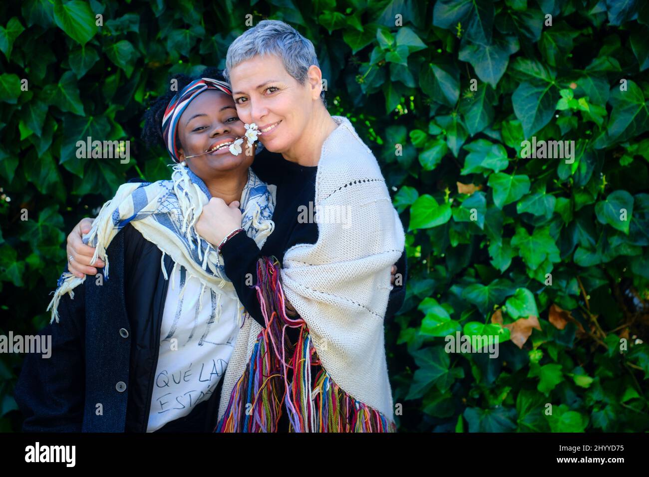 Reife kaukasische Frau und afrikanische junge Frau umarmen sich in einem Garten mit einem Laubhintergrund. Stockfoto