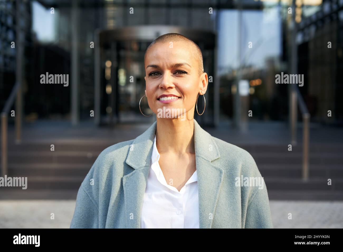 Fröhliche Frau mittleren Alters mit rasierten Haaren und Blick auf die Kamera. Geschäftsleute im Freien auf dem Hintergrund mit Gebäuden. Corporate. Stockfoto
