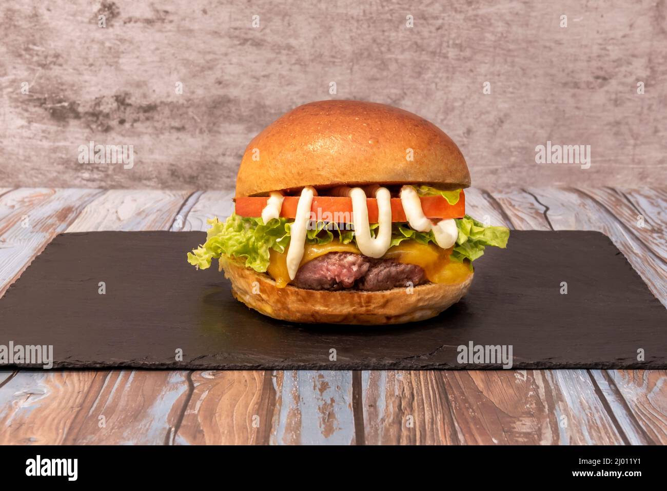 Klassischer Rinderkäseburger mit Tomaten, Eisbergsalat, Mayonnaise und Cheddar-Käse auf einem schwarzen Schieferbrett Stockfoto