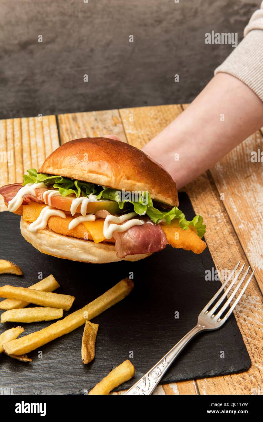 Eine Hand, die einen knusprigen Hähnchenburger mit frisch geschnittenen Tomaten, frischem Salat und Mayonnaise auf einem schwarzen Schieferteller mit pommes frites und einem fest hält Stockfoto