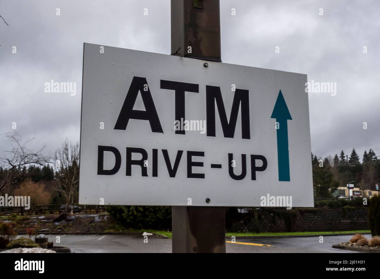 Leicht abgewinkelte Ansicht eines weißen ATM-Drive-Up-Schilds mit einem Richtungspfeil, der auf den Beginn des Drive-Thru zeigt und gegen einen bewölkten Himmel geschossen ist Stockfoto