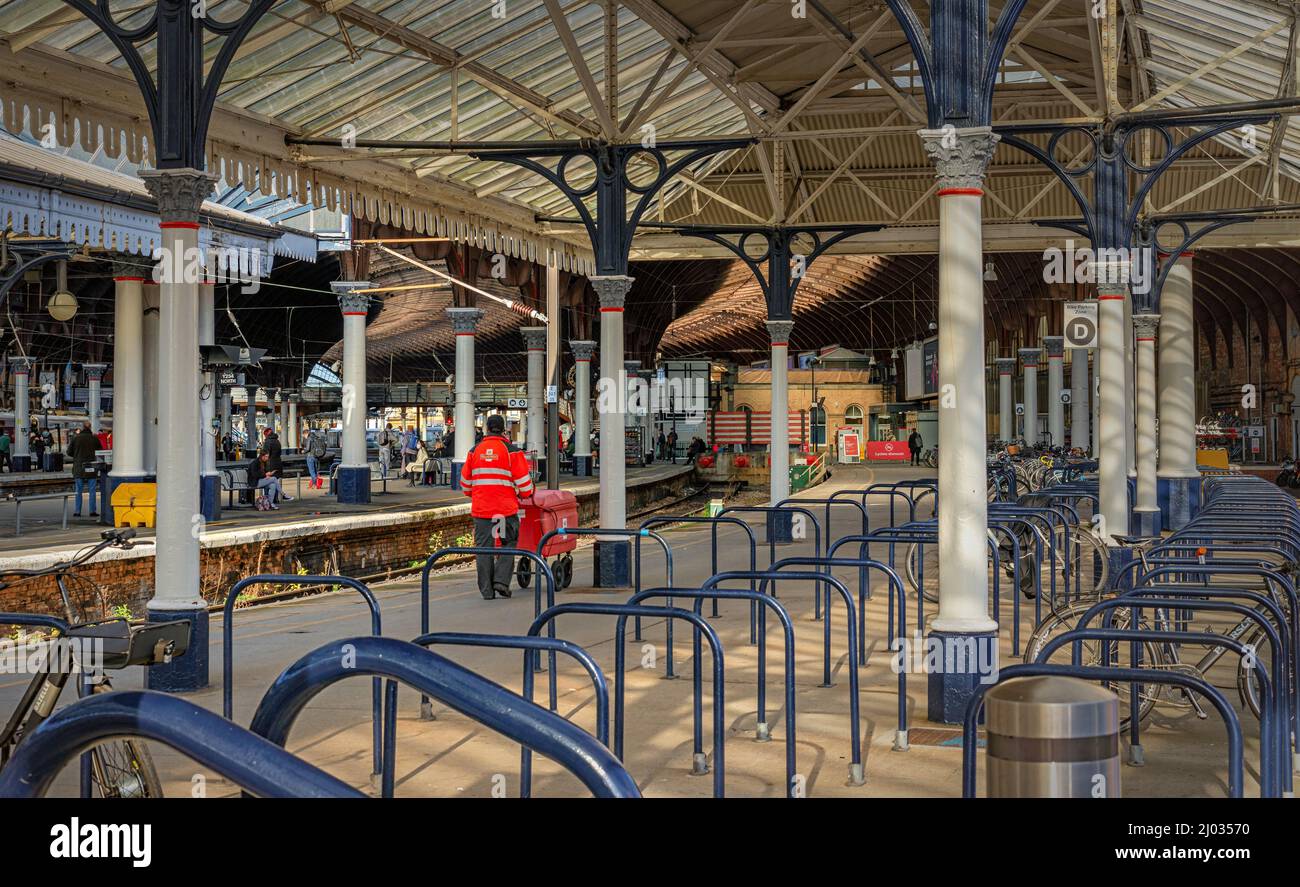 Ein Postbote schiebt einen Trolley entlang einer Bahnhofsplattform. Ein historisches Vordach aus dem 19.. Jahrhundert ist über dem Kopf und Fahrradständer stehen im Vordergrund. Stockfoto