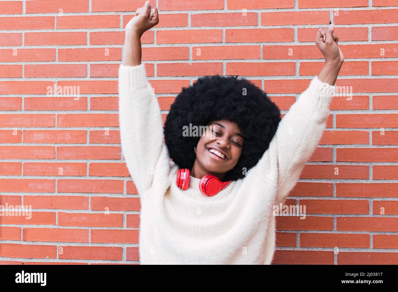 Fröhliche schwarze Frau mit roten Kopfhörern am Hals, Afro-Frisur und perfektem weißen Lächeln tanzen, Spaß haben und lächeln mit geschlossenen Augen. Speicherplatz kopieren Stockfoto