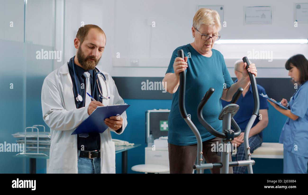 Arzt gibt der älteren Frau auf einem stationären Fahrrad Ratschläge und verwendet alternative Medizin zur Behandlung von Körperverletzungen. Facharzt, der den Patienten bei der Genesung unterstützt und die Mobilität erhöht. Stockfoto