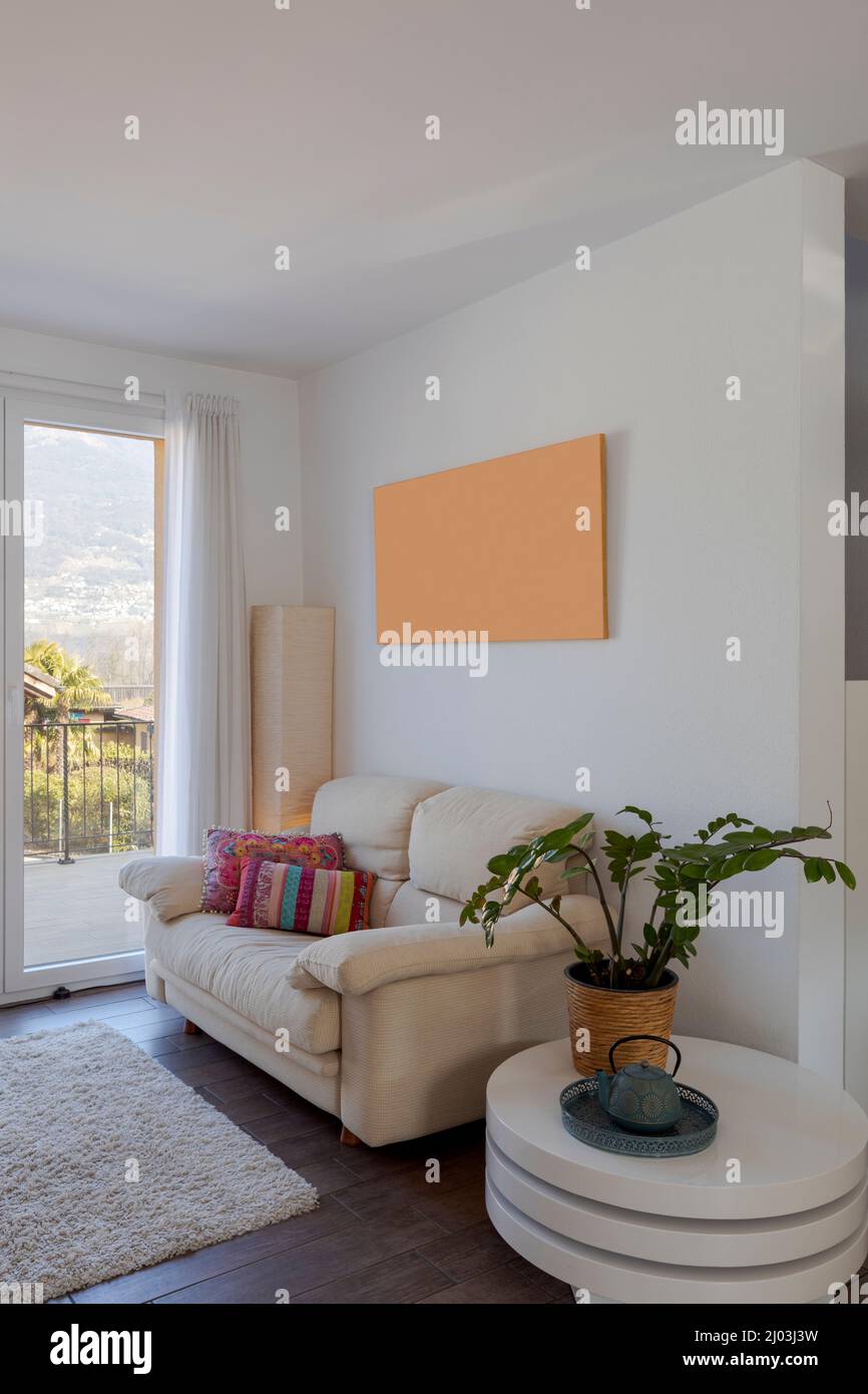 Kleines Wohnzimmer mit Sofa und bunten Kissen. Es gibt einen kleinen Tisch mit einer Pflanze darauf. Fenster mit Blick auf die Natur. Niemand drinnen Stockfoto