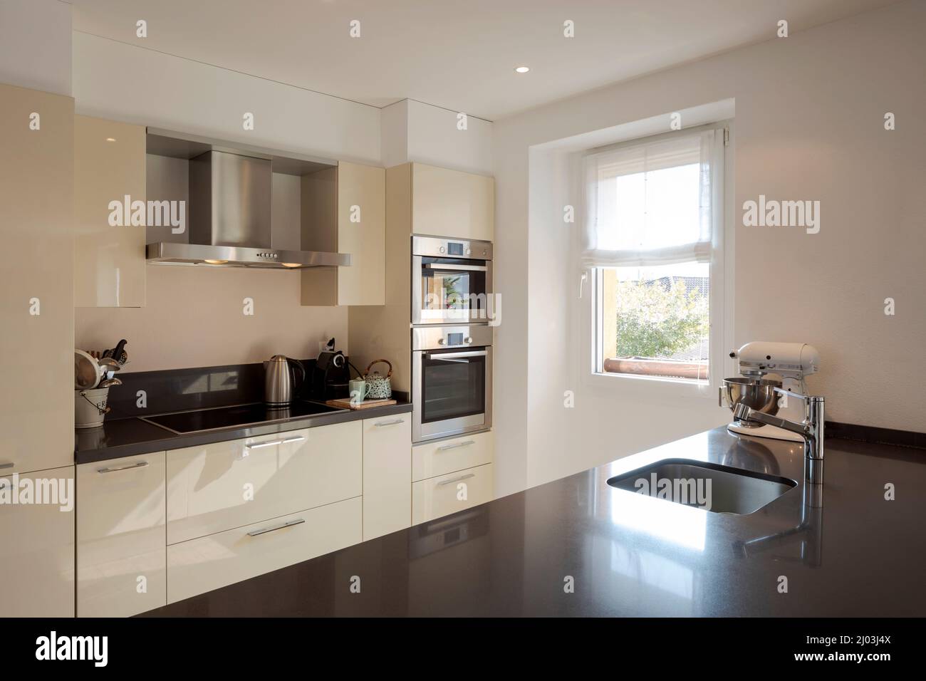 Moderne Küchenausstattung mit großer dunkler Insel mit Hockern. Waschbecken aus Edelstahl und helles Fenster. Stockfoto