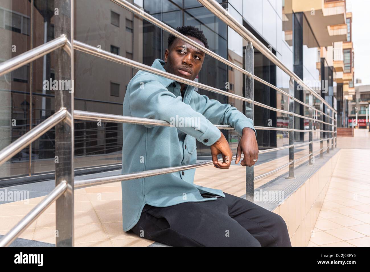 Seriöser junger afroamerikanischer Tausendjähriger Mann mit dunklem lockigem Haar in stilvollem Outfit, das sich auf Metallgeländer stützt und beim Sitzen nachdenklich davonschaut Stockfoto