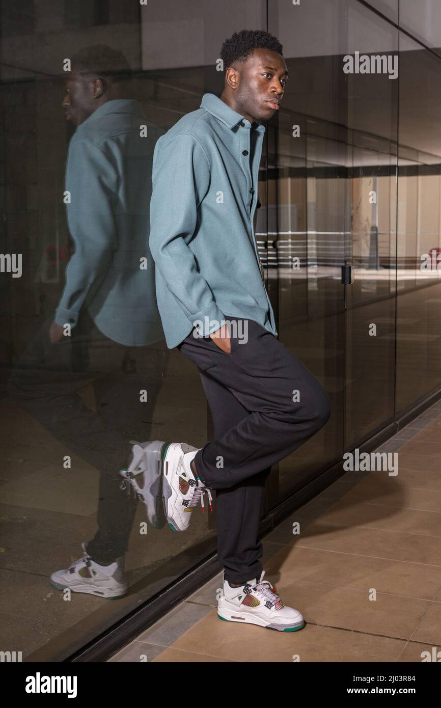 Seitenansicht nachdenklicher junger afroamerikanischer Mann in trendigem Outfit und Sneakers, die an einer Glaswand im modernen Gebäudekorridor gelehnt sind und wegschauen Stockfoto