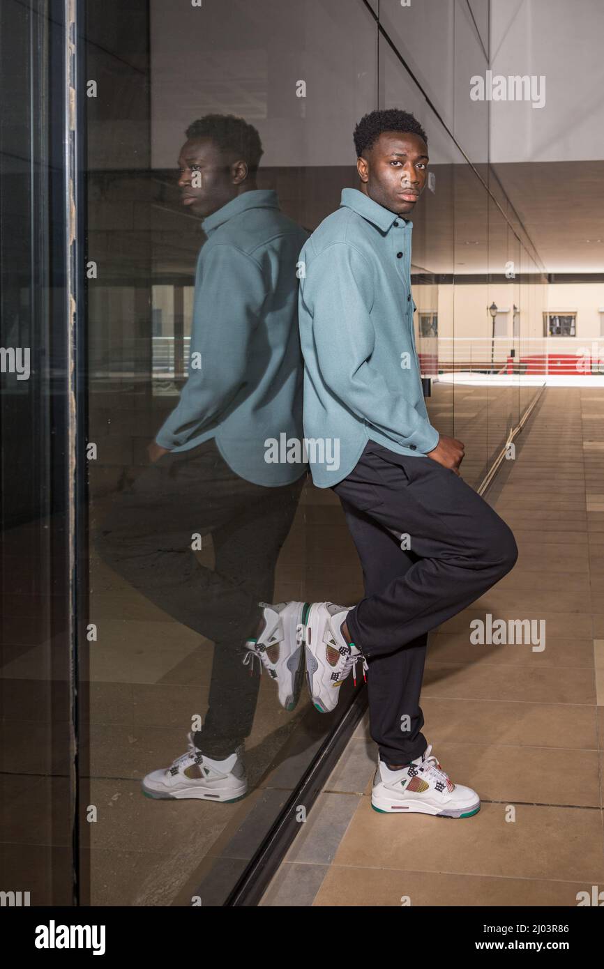 Seitenansicht Ganzkörper aus selbstsicherem jungen schwarzen männlichen Millennial mit dunklem Haar in trendigem Outfit und Sneakers, die in Passage und Look an einer Glaswand angelehnt sind Stockfoto