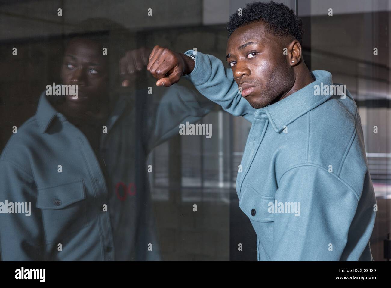 Seitenansicht eines selbstbewussten jungen afroamerikanischen Mannes mit lockig dunklem Haar in stilvoller Jacke, die an einer Glaswand lehnt und in einem modernen Gebäude die Kamera anschaut Stockfoto
