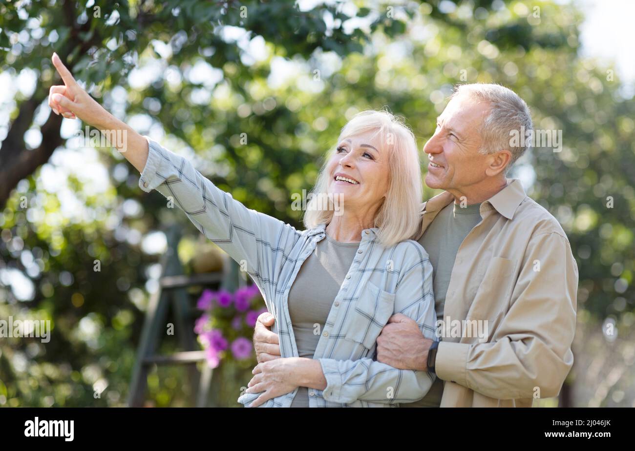 Glückliches Rentnerpaar, das im Garten Zeit zusammen genießt, am Frühlingstag spazierengeht, umarmt und eine Frau etwas zeigt Stockfoto