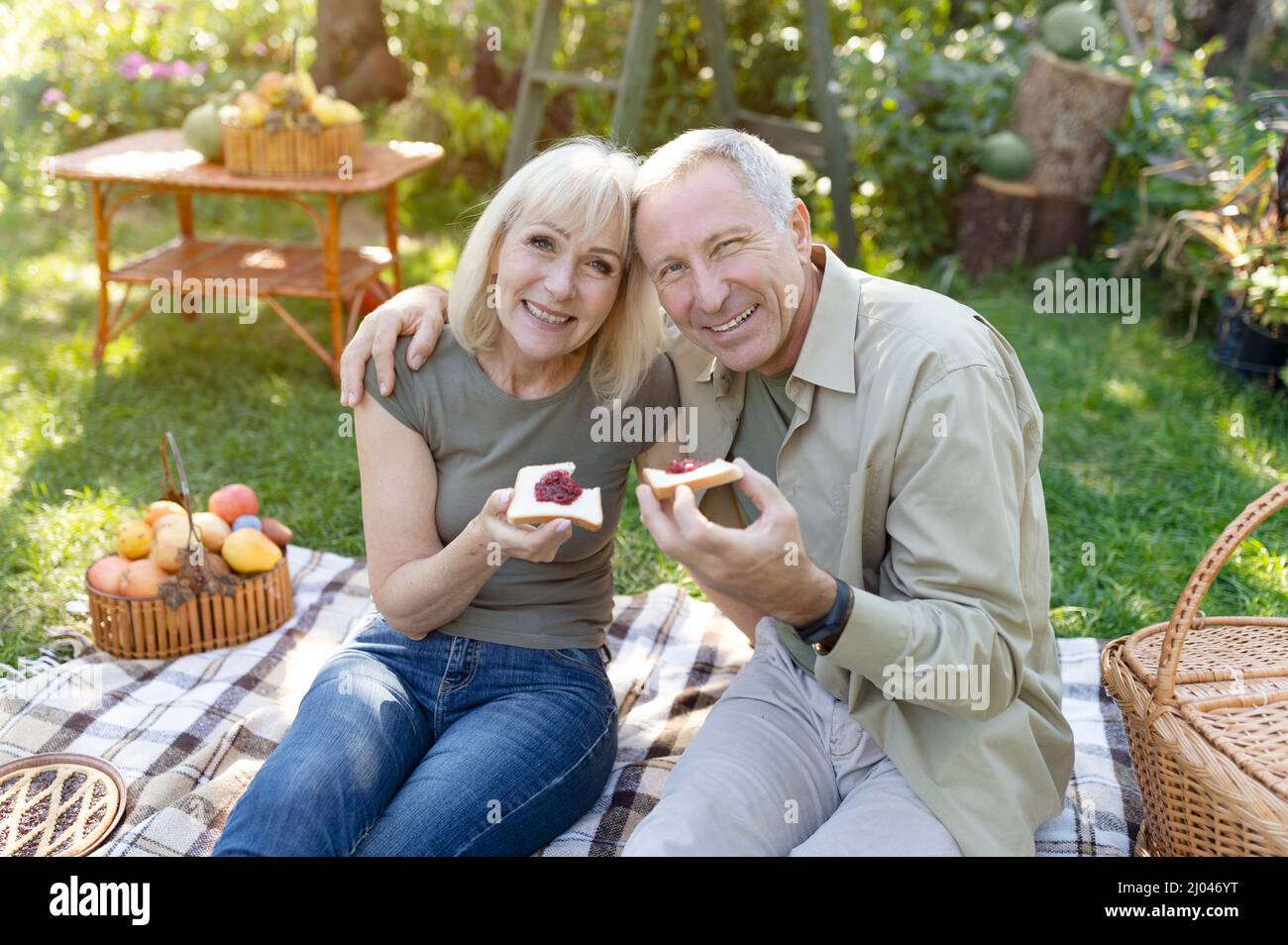 Liebende ältere Ehepartner, die Picknick machen, auf der Decke im Garten sitzen und mit Marmelade Toast essen und die Kamera lächeln Stockfoto