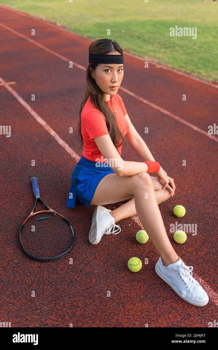 Asiatische junge hübsche Frau mit schwarzem Stirnband sitzt auf dem Sportstadion mit dem Tennisschläger und Kugeln. Sport im Freien. Hochwertige Fotos Stockfoto
