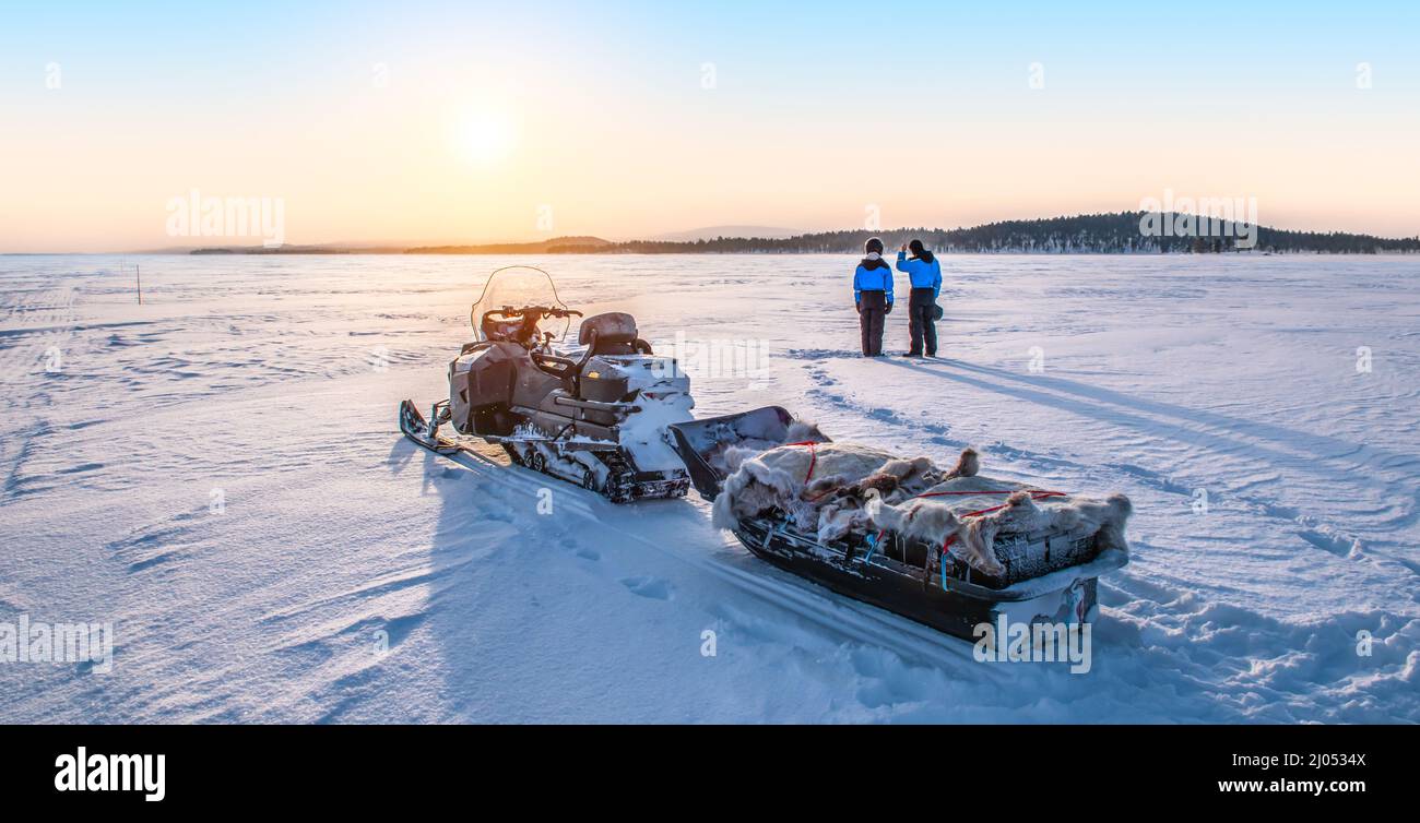 Das junge Paar genießt den Sonnenaufgang in der Nähe des Schneemobiles auf dem gefrorenen See Inari in Finnland, Lappland. Stockfoto