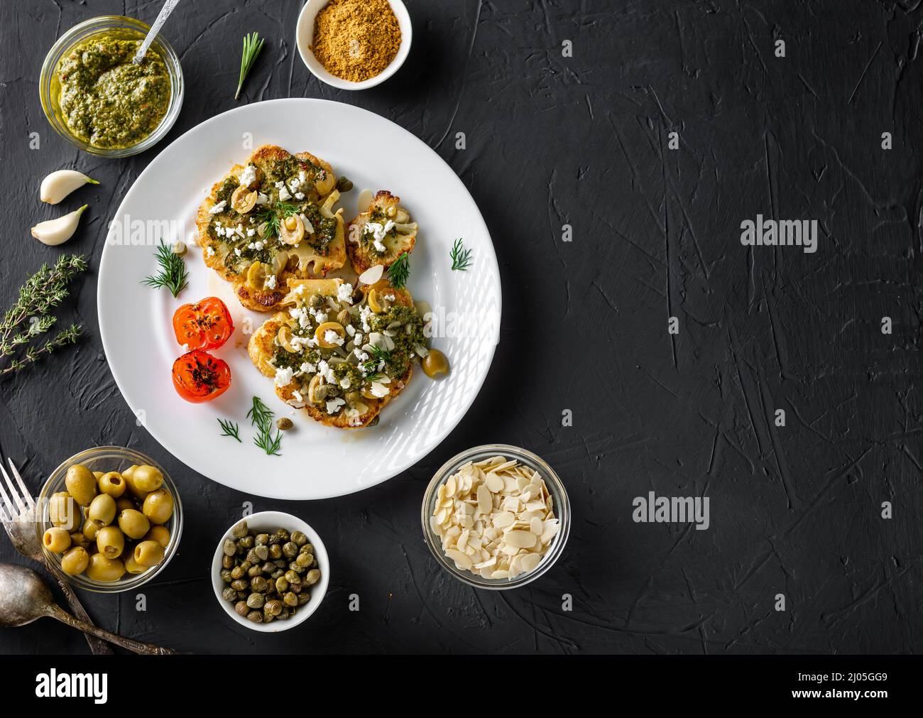 Blumenkohlesteak mit Gewürzen, Chimichurri-Sauce, Mandelflocken, Oliven, gebratenen Kirschtomaten und Kapern auf einem weißen Teller. Dunkler Hintergrund. Copyspace Stockfoto