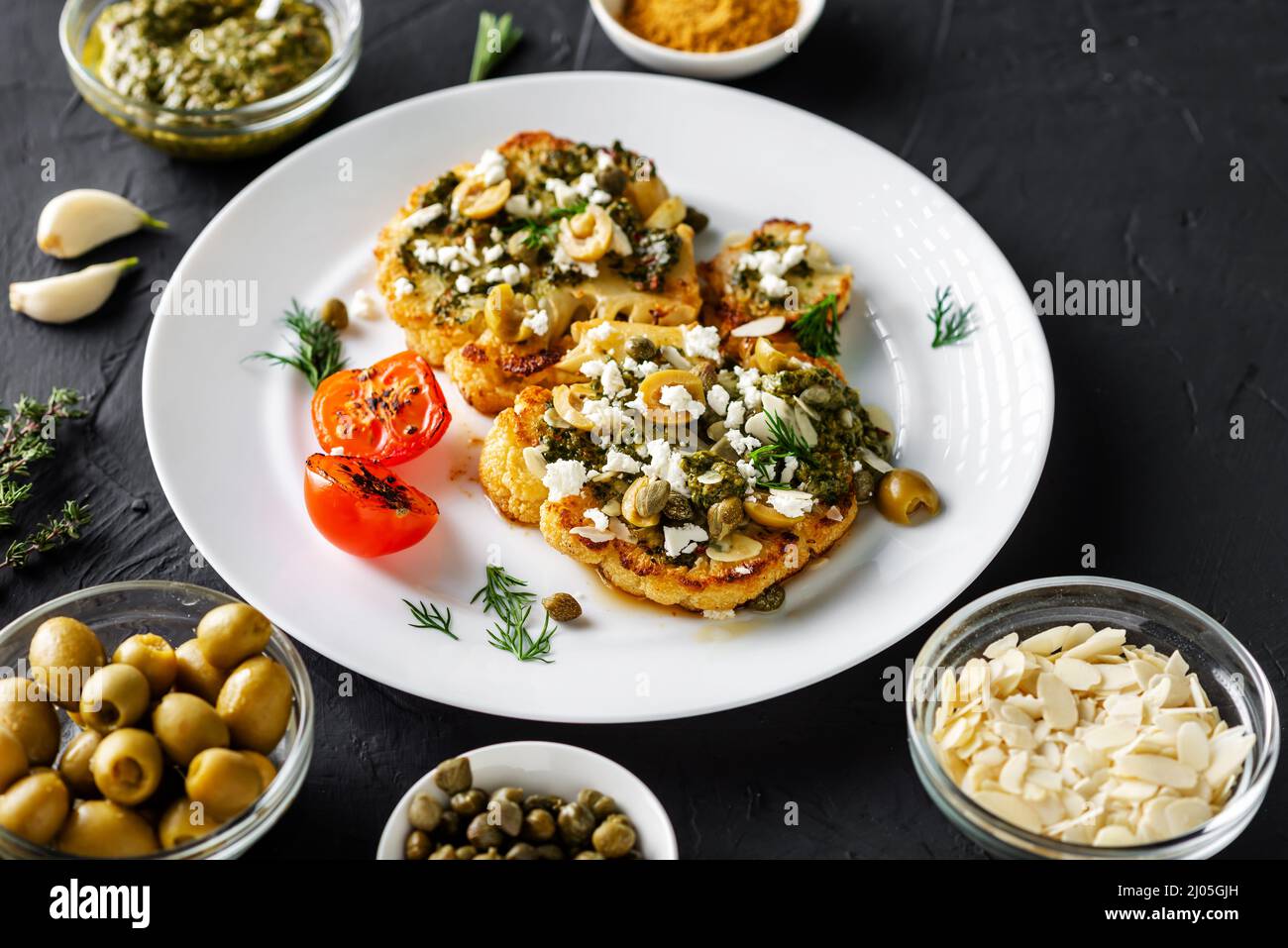 Blumenkohlesteak mit Gewürzen, Chimichurri-Sauce, Mandelflocken, Oliven, gebratenen Kirschtomaten und Kapern auf einem weißen Teller. Dunkler Hintergrund. Stockfoto