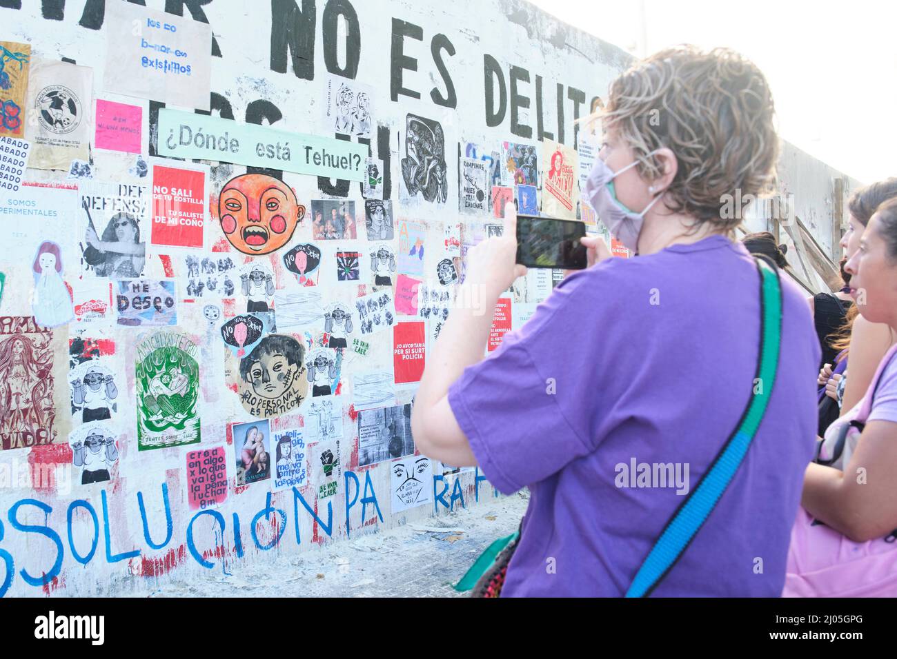 CABA, Buenos Aires, Argentinien; 8. März 2022: Internationaler feministischer Streik. Frau mit einem violetten Hemd, die Fotos von einer Wand voller Poster und f Stockfoto