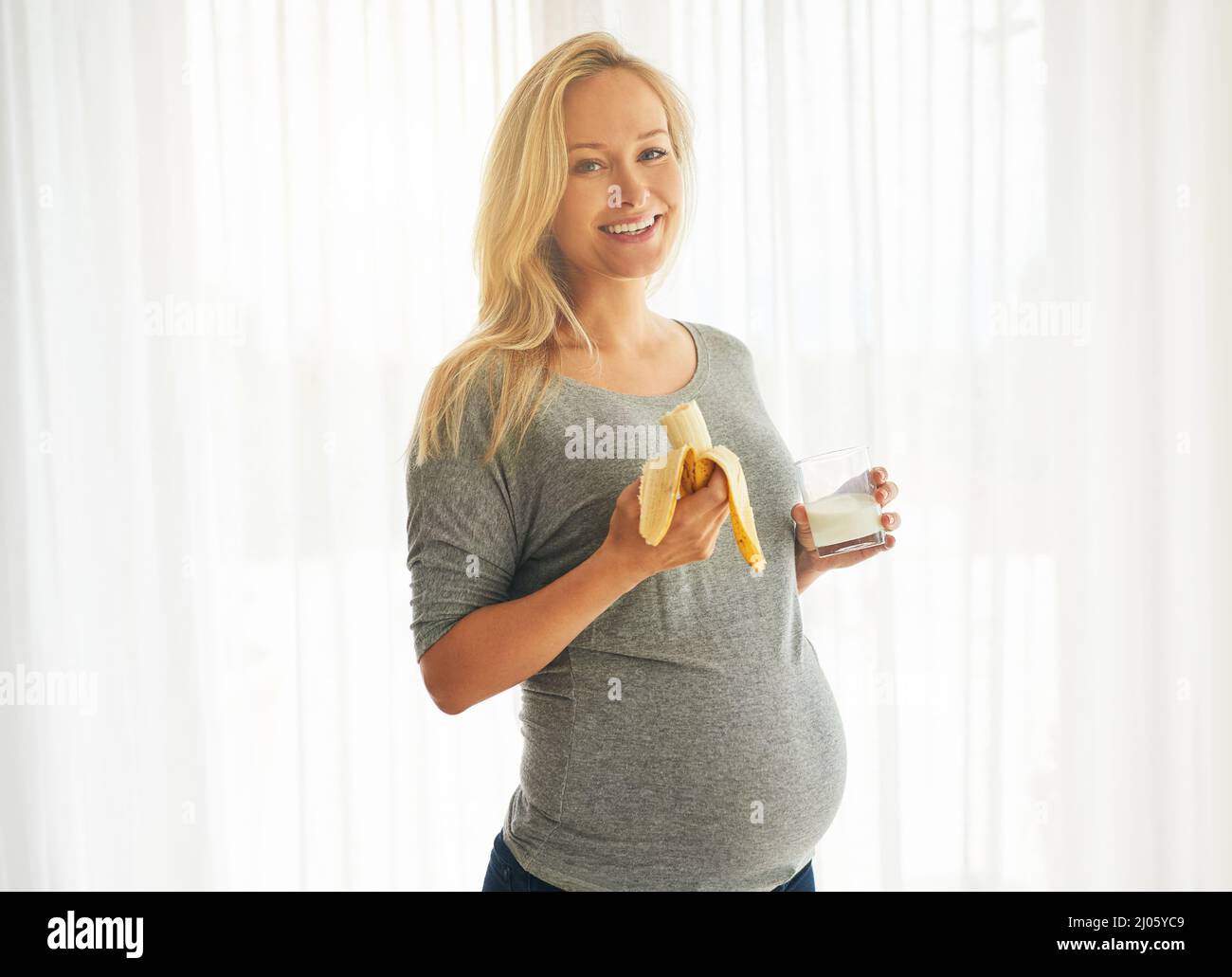 Gesunde Mama, gesundes Baby. Porträt einer glücklichen Schwangeren, die zu Hause eine Banane isst und ein Glas Milch trinkt. Stockfoto