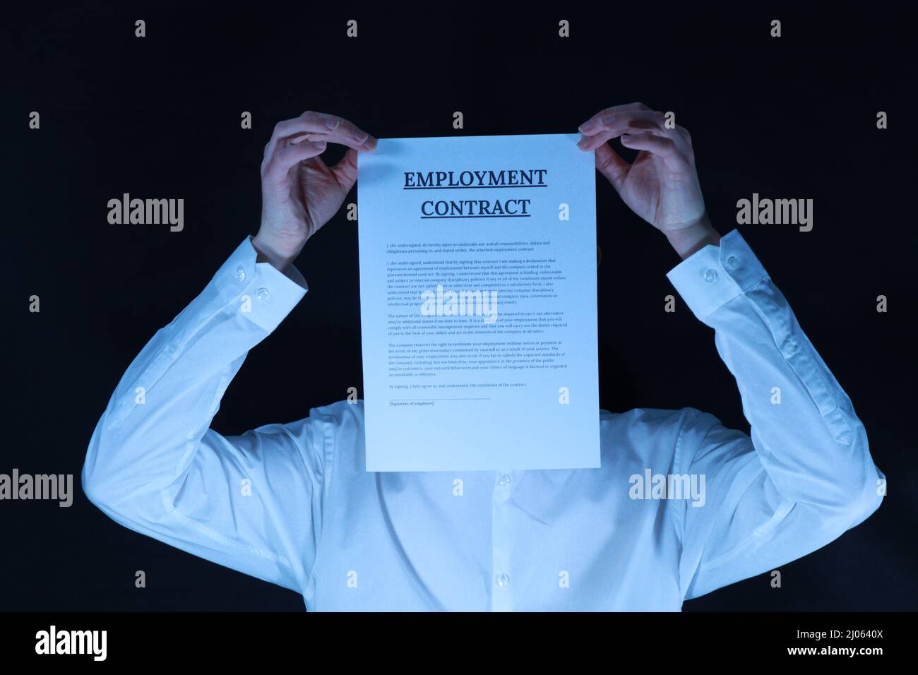 Der gesichtslose Mitarbeiter - Ein Mann in einem weißen Hemd, der vor einem schwarzen Hintergrund steht, hält einen Mitarbeitervertrag über seinem Gesicht Stockfoto