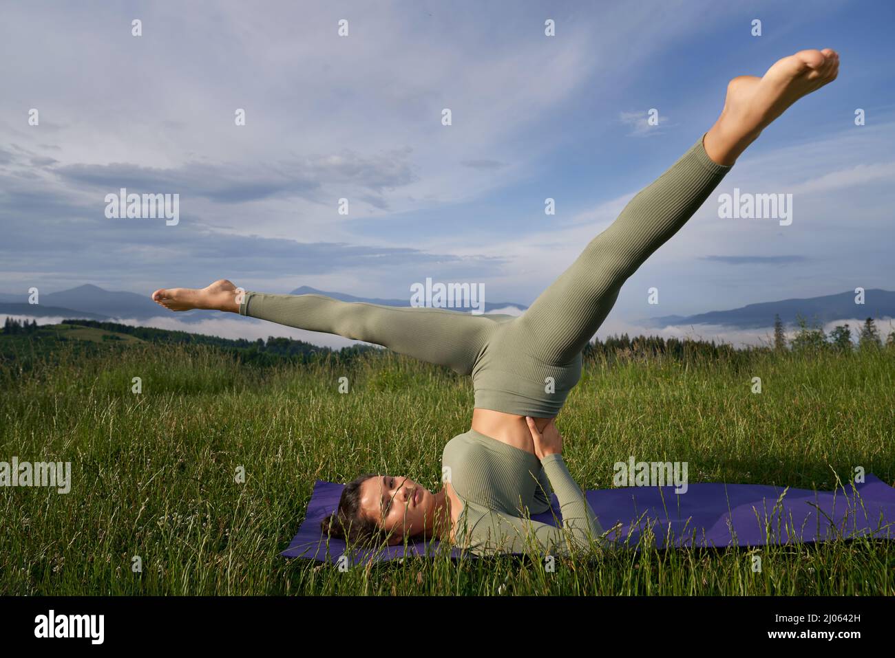Junge aktive Frau in Sportkleidung, die morgens auf einer Yogamatte im Freien akrobatische Übungen macht. Tolle Aussicht auf grüne Berge im Hintergrund. Stockfoto
