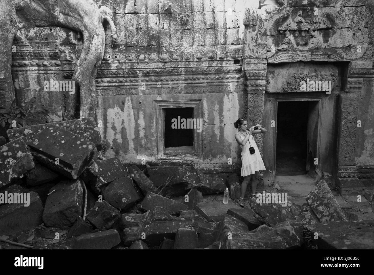 Eine Besucherin, die mit ihrer Handykamera zwischen den Ruinen des Ta Prohm Tempels in Siem Reap, Kambodscha, fotografiert (schwarz-weiße Version). Einst als Drehort für einen Hollywood-Film von 2001 verwendet Lara Croft: Tomb Raider mit Angelina Jolie und vor allem bekannt für seine riesigen Wurzeln aus wilden Bäumen, die tief zwischen den Steinen verwurzelt sind, war Ta Prohm ein Kloster für Studenten des Mahayana-Buddhismus, bevor es verlassen und für Hunderte von Jahren vergessen wurde. Stockfoto