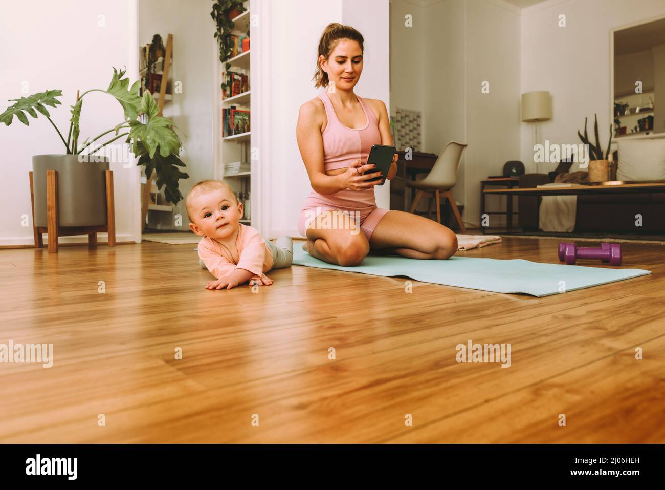 Gesunde Mutter nutzt die Fitness-App, während sie mit ihrem Baby trainiert. Yogi-Mama hält ein Smartphone, während sie auf einer Übungsmatte kniet. Neue Mutter folgt ihr Stockfoto