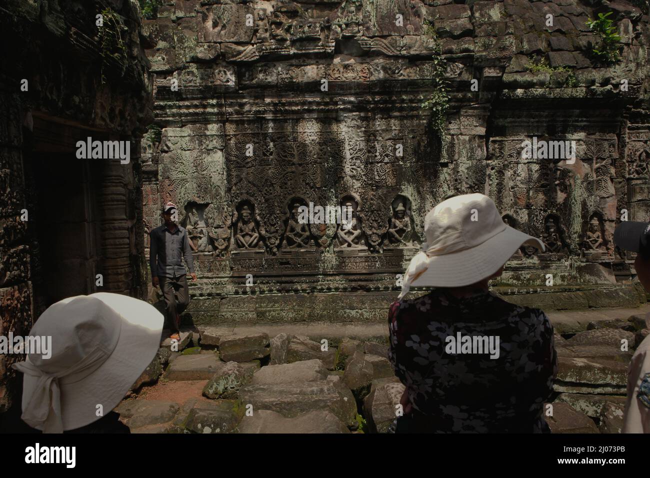 Frauen, die stehen, während sie einem Reiseleiter zuhören, der Ihnen etwas erklärt, und überblicken eine verwitterte Wand, die mit Basreliefs dekoriert ist, im Preah Khan Tempel in Siem Reap, Kambodscha. Stockfoto